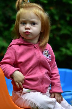 Açık havada turuncu bir sandalyede oturan, pembe kapüşonlu beyaz pantolon giyen, güneşli bir günde kameraya bakarken neşeli bir şekilde somurtan sevimli kızıl saçlı çocuk..