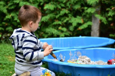 Çizgili kapüşonlu küçük çocuk mavi bir kum havuzunun yanında oturuyor ve dışarıda kum oyuncaklarıyla oynuyor, bahçedeki sakin ve güneşli bir günde aktivitelerine odaklanıyor..