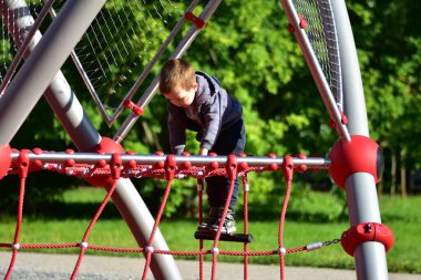 Çocuk çocuk oyun parkında kırmızı ip yapısına tırmanıyor, hareket ve koordinasyon üzerine yoğunlaşıyor. Aktif açık hava oyunu güç ve cesaret kazanıyor..