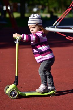 Kırmızı oyun alanı yakınlarında yeşil scooter kullanan küçük bir kız. Güneşli bir sabahta enerjik oyun ve koordinasyon eğitimi.
