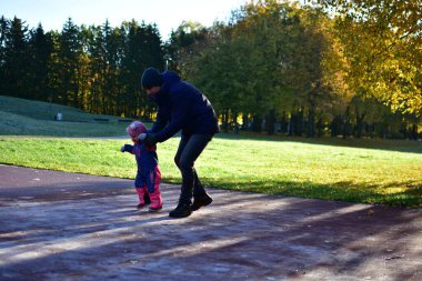 Baba, küçük kızın sonbahar parkında buzlu oyun parkında yürümesine yardım ediyor. Sıcak güneş ışığı ve renkli ağaçlar dışarıda rahat bir aile havası yaratıyor. Babası ve küçük kızı soğuk sonbaharda dışarıda oynuyorlar.