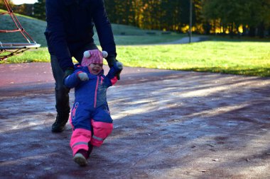 Kış tulumlarıyla gülen küçük kız güneşli bir sonbahar sabahında ebeveynleriyle birlikte buzlu oyun alanında yürümeyi öğreniyor. Neşeli açık hava aile anı.