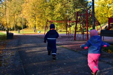Güneşli bir sonbahar gününde salıncaklarla ve tırmanma çerçeveleriyle çocuk parkına doğru koşan sıcak tulumlu iki çocuk, aktif çocukluk anı. Erkek ve kız kardeş boş sonbahar parkında birlikte oynuyorlar.