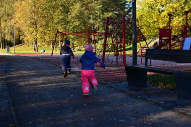 Güneşli bir sonbahar gününde salıncaklarla ve tırmanma çerçeveleriyle çocuk parkına doğru koşan sıcak tulumlu iki çocuk, aktif çocukluk anı. Erkek ve kız kardeş boş sonbahar parkında birlikte oynuyorlar.