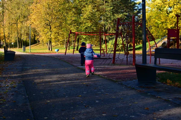 Güneşli bir sonbahar gününde salıncaklarla ve tırmanma çerçeveleriyle çocuk parkına doğru koşan sıcak tulumlu iki çocuk, aktif çocukluk anı. Erkek ve kız kardeş boş sonbahar parkında birlikte oynuyorlar.