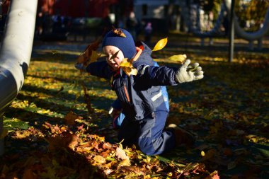Mavi kar kıyafetli gülümseyen çocuk güz yapraklarını havaya fırlatıyor, güneşli bir sonbahar akşamında kız kardeşinin yanında oynuyor kahkahalarla dolu..