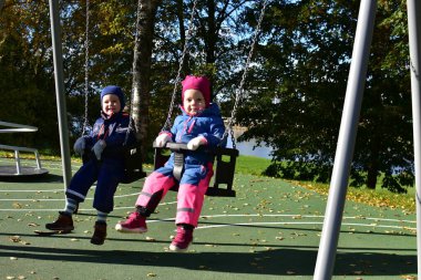Renkli kış tulumları içinde iki neşeli çocuk güneşli bir sonbahar bahçesinde ağaçlar ve yapraklarla çevrili bir şekilde sallanarak açık havada serin sonbahar havasının keyfini çıkarıyorlar..