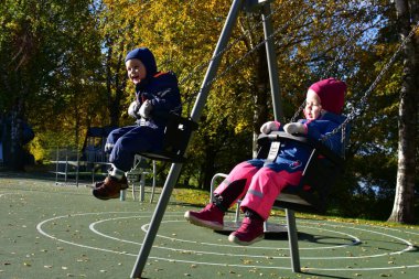 Renkli kış tulumları içinde iki neşeli çocuk güneşli bir sonbahar bahçesinde ağaçlar ve yapraklarla çevrili bir şekilde sallanarak açık havada serin sonbahar havasının keyfini çıkarıyorlar..