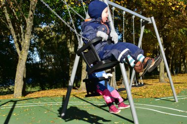 Renkli kış tulumları içinde iki neşeli çocuk güneşli bir sonbahar bahçesinde ağaçlar ve yapraklarla çevrili bir şekilde sallanarak açık havada serin sonbahar havasının keyfini çıkarıyorlar..