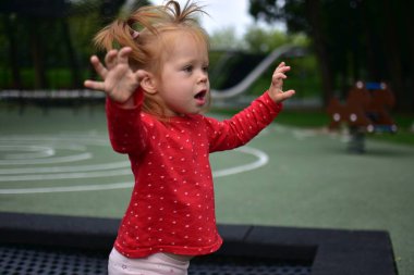 Küçük kız trambolinde oynarken kollarını kaldırıyor, kırmızı tişört, şaşkın ifade yüz, yeşil park ortamında eğlenceli dışarıda oyun zamanı.