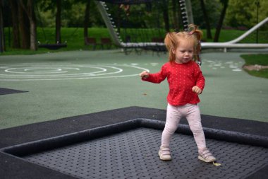 Trambolin yüzeyini keşfeden küçük bir kız, kırmızı gömlek ve taytlarla yürüyor ve oynuyor, modern parkta açık havada eğlenceli bir çocukluk anı..
