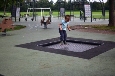 Çocuk, elinde tüy tutarak çocuk parkının içinde trambolinde zıplıyor. Yeşil park çevresindeki doğa hareketi ve enerji anı..