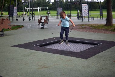 Çocuk, elinde tüy tutarak çocuk parkının içinde trambolinde zıplıyor. Yeşil park çevresindeki doğa hareketi ve enerji anı..