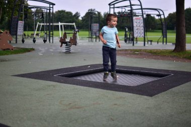 Çocuk, elinde tüy tutarak çocuk parkının içinde trambolinde zıplıyor. Yeşil park çevresindeki doğa hareketi ve enerji anı..