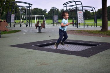 Çocuk, elinde tüy tutarak çocuk parkının içinde trambolinde zıplıyor. Yeşil park çevresindeki doğa hareketi ve enerji anı..