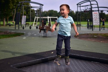 Çocuk, elinde tüy tutarak çocuk parkının içinde trambolinde zıplıyor. Yeşil park çevresindeki doğa hareketi ve enerji anı..