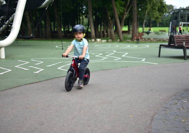 Parktaki oyun parkında kaskla bisiklete binen küçük çocuk, dışarıda güvenli bisiklet sürme anı koordinasyon becerilerini ve doğada aktif yaşam tarzını geliştiriyor..