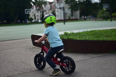 Kask takan küçük çocuk parkta asfalt yolda kırmızı denge bisikletini itiyor, dışarıda yeşil ortamda dengeyi ve hareketi öğreniyor. Kimliği belirsiz çocuk yok..