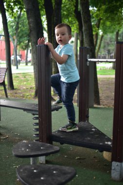 Parkta modern oyun alanı yapısına tırmanan çocuk, korkulukları tutuyor, odaklanmış ifade, günlük kıyafetler ve spor ayakkabıları, yeşil doğa arka planı. Etkin oyun ve fiziksel gelişim anı.