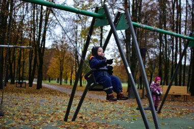 Küçük çocuk serin bir sonbahar günü, renkli altın yapraklar ve ağaçlarla çevrili bir şekilde oyun alanında sallanmaktan hoşlanıyor. Samimi açık hava çocukluk anı eğlence ve neşe dolu.