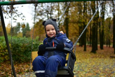 Tatlı çocuk, sonbaharda parkta nazikçe sallanarak eğleniyor. Soğuk bir gün için sıcak bir şekilde gülümsüyor ve giyiniyor. Ağaçlar ve düşen yapraklar arasında sakin ve tatlı bir çocukluk anı..