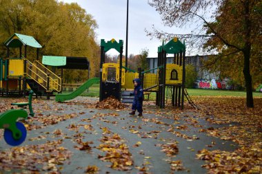 Sonbahar çocuk parkını keşfeden ve düşen yaprakların arasında yürüyen sıcacık giysiler giyen bir çocuk. Mevsimsel açık hava çocukluk anı. Renkli doğa ve oyun yapılarıyla çevrili..