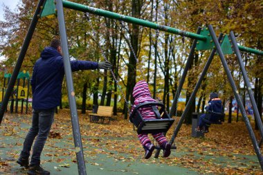 Başka bir çocuk sonbahar parkında sallanırken ebeveyn çocuğu salıncakta sallıyor. Eğlenceli, temiz hava ve renkli yapraklarla dolu açık hava aile aktiviteleri..