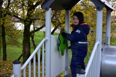 Mavi elbiseli küçük bir çocuk, sonbahar günü oyun sahasında yeşil dönen tekerlekle oynuyor, mutlu bir gülümseme ve neşeli bir ifade, açık havada öğrenme ve güzel motor beceriler geliştirme..