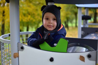 Koyu mavi kar giysili ve kulakları olan küçük bir çocuk sonbahar parkında modern oyun alanında oynuyor, çocukluk, öğrenme ve açık hava eğlencesi kavramını keşfediyor ve bakıyor..