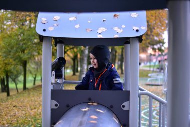 Mavi kar kıyafetli ve şapkalı küçük çocuk teleskopla oyun kulesinde oynuyor, gülümsüyor ve izliyormuş gibi yapıyor, sonbahar parkı arka planı sarı yapraklar, hayal gücü ve eğlenceyle dolu..