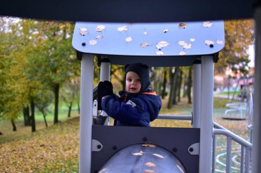 Mavi kar kıyafetli ve şapkalı küçük çocuk teleskopla oyun kulesinde oynuyor, gülümsüyor ve izliyormuş gibi yapıyor, sonbahar parkı arka planı sarı yapraklar, hayal gücü ve eğlenceyle dolu..