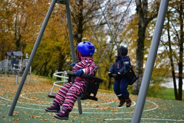 Ilık kar kıyafetleri giymiş iki küçük çocuk, sonbahar parkında bir oyun bahçesinde sallanıyorlar, yerde renkli yapraklar, dengeyi, koordinasyonu ve eğlenceyi destekleyen neşeli açık hava aktiviteleri..