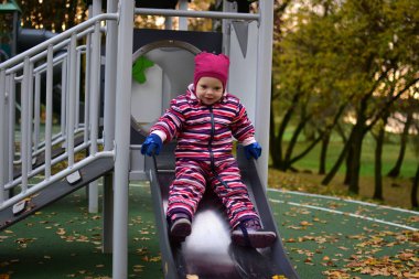 Pembe çizgili kar elbisesi içinde gülümseyen çocuk oyun parkından kayarak kayıyor, sarı yapraklar, eğlence anlayışı, çocukluk ve açık hava aktiviteleriyle dolu sonbahar oyun zamanının tadını çıkarıyor..