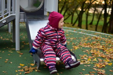 Pembe çizgili kar elbisesi içinde gülümseyen çocuk oyun parkından kayarak kayıyor, sarı yapraklar, eğlence anlayışı, çocukluk ve açık hava aktiviteleriyle dolu sonbahar oyun zamanının tadını çıkarıyor..
