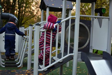 Renkli kar giysileri içinde iki çocuk, serin bir sonbahar öğleden sonra tırmanarak ve oyun alanı yapısını keşfederken, takım çalışması ve merak, açık havada koordinasyon ve denge becerilerini geliştirirken,.