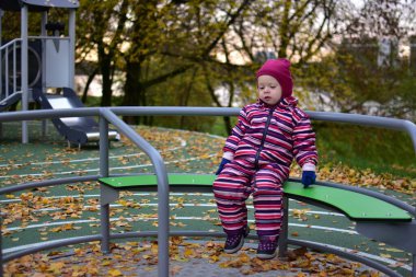 Pembe çizgili kar elbisesiyle gülümseyen çocuk oyun sahasında sonbahar yapraklarıyla kaplı bir şekilde döner kavşakta oturuyor, nehir kenarındaki parkta eğleniyor, çocukluk mutluluğu ve hareketinin keyfini çıkarıyor..