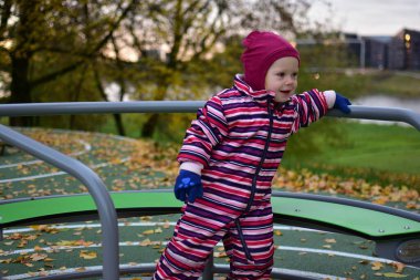 Pembe çizgili kar elbisesi içinde gülen çocuk oyun sahasında sonbahar yapraklarıyla kaplı döner kavşakta oynuyor, nehir kenarındaki parkta eğleniyor, çocukluk mutluluğu ve hareketinin keyfini çıkarıyor..