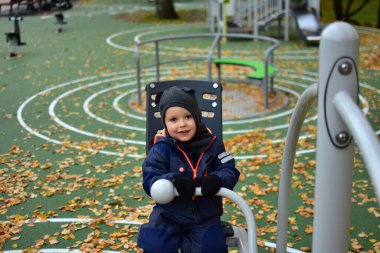 Mavi kar kıyafetli, gülen anaokulu çocuğu, sonbahar yapraklarıyla kaplı bahçedeki spor salonunda oturmuş, serin bir sonbahar gününde fiziksel aktivitenin ve temiz havanın tadını çıkarıyor..