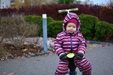 Sonbaharda açık hava parkında küçük dengeli bir bisikletin üzerinde oturan çizgili bir kar elbisesi içindeki çocuk. Yumuşak renkler, çalılar ve sakin bir ifade basit bir çocukluk anını yakalar..