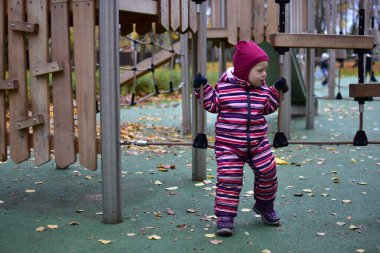 Çizgili bir kar elbisesi içinde, oyun parkında ahşap bir ip köprüyü keşfeden bir çocuk. Sonbahar yaprakları ve yeşil zemin hareketlilik ve merakla dolu canlı bir açık hava ortamı yaratır..