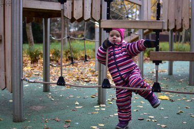 Çizgili bir kar elbisesi içinde, oyun parkında ahşap bir ip köprüyü keşfeden bir çocuk. Sonbahar yaprakları ve yeşil zemin hareketlilik ve merakla dolu canlı bir açık hava ortamı yaratır..