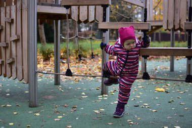 Çizgili bir kar elbisesi içinde, oyun parkında ahşap bir ip köprüyü keşfeden bir çocuk. Sonbahar yaprakları ve yeşil zemin hareketlilik ve merakla dolu canlı bir açık hava ortamı yaratır..