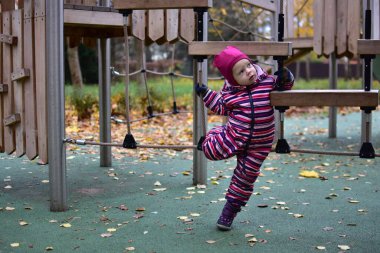 Çizgili bir kar elbisesi içinde, oyun parkında ahşap bir ip köprüyü keşfeden bir çocuk. Sonbahar yaprakları ve yeşil zemin hareketlilik ve merakla dolu canlı bir açık hava ortamı yaratır..