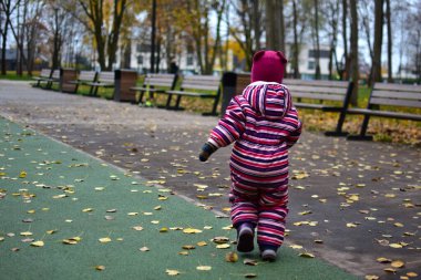 Çizgili bir kar elbisesi içinde, sonbahar yapraklarıyla kaplı bir parkta yürüyen anonim bir çocuk. Banklar, ağaçlar ve yumuşak ışık mevsimsel renklerle dolu huzurlu bir çocukluk sahnesi yaratır..