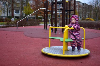 Sonbahar boyunca açık hava oyun parkında sarı bir atlıkarınca üzerinde oturan çizgili bir kar elbisesi içindeki çocuk. Ağaçlı ve yumuşak kauçuk döşemeli şehir parkı her gün sakin bir çocukluk anı yaratır..