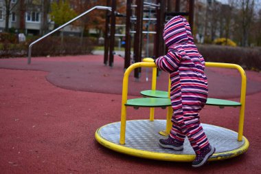 Çizgili bir kar elbisesi içinde, sonbahar boyunca açık hava oyun parkında sarı bir atlıkarıncada dönen bir bebek. Ağaçlı ve yumuşak kauçuk döşemeli şehir parkı her gün sakin bir çocukluk anı yaratır..