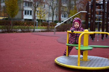 Çizgili bir kar elbisesi içinde, sonbahar boyunca açık hava oyun parkında sarı bir atlıkarıncada dönen bir bebek. Ağaçlı ve yumuşak kauçuk döşemeli şehir parkı her gün sakin bir çocukluk anı yaratır..