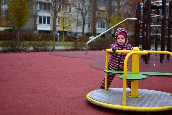 Çizgili bir kar elbisesi içinde, sonbahar boyunca açık hava oyun parkında sarı bir atlıkarıncada dönen bir bebek. Ağaçlı ve yumuşak kauçuk döşemeli şehir parkı her gün sakin bir çocukluk anı yaratır..