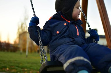 Mavi kışlık takım elbiseli, salıncakta oturan ve yan tarafa bakarken gülümseyen bir çocuğun yakın çekimi. Soğuk havada oynamak için samimi bir an..