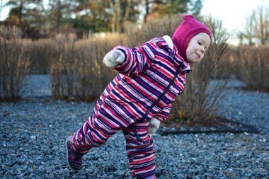 Çizgili kışlık takım elbiseli bir kız dışarıda çakıllı bir yolda neşeyle koşuyor ya da zıplıyor. Aktif çocukluk hareketinin neşeli samimi anı.